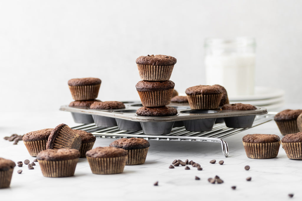 mini grain-free brownie bites in a mini muffin tin, with 3 brownies stacked above the others. More brownie bites and chocolate chips on counter, and glass of milk in background