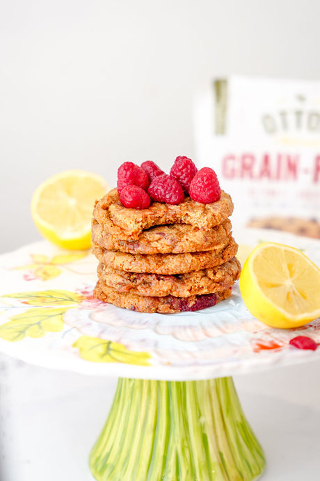 5 stacked grain-free lemon curd raspberry cookies, surrounded by lemon halves and topped with fresh raspberries. Bag of Otto's Naturals Ultimate Grain-Free Cookie Mix in background