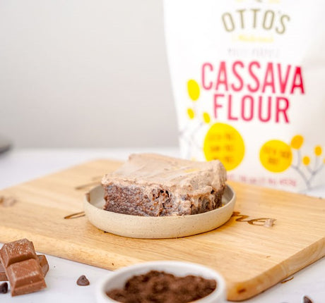 Grain-Free Chocolate Tres Leches Cake Slice sitting on plate on cutting board. Bag of Otto's Naturals Cassava Flour in background