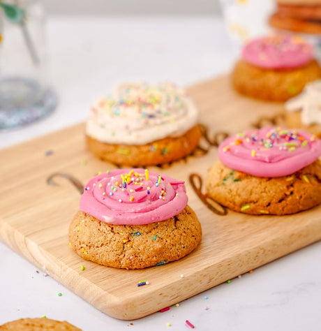 Crumbl Copycat cookies (grain-free) sitting on cutting board. With pink frosting and white frosting and rainbow sprinkles for confetti cookies