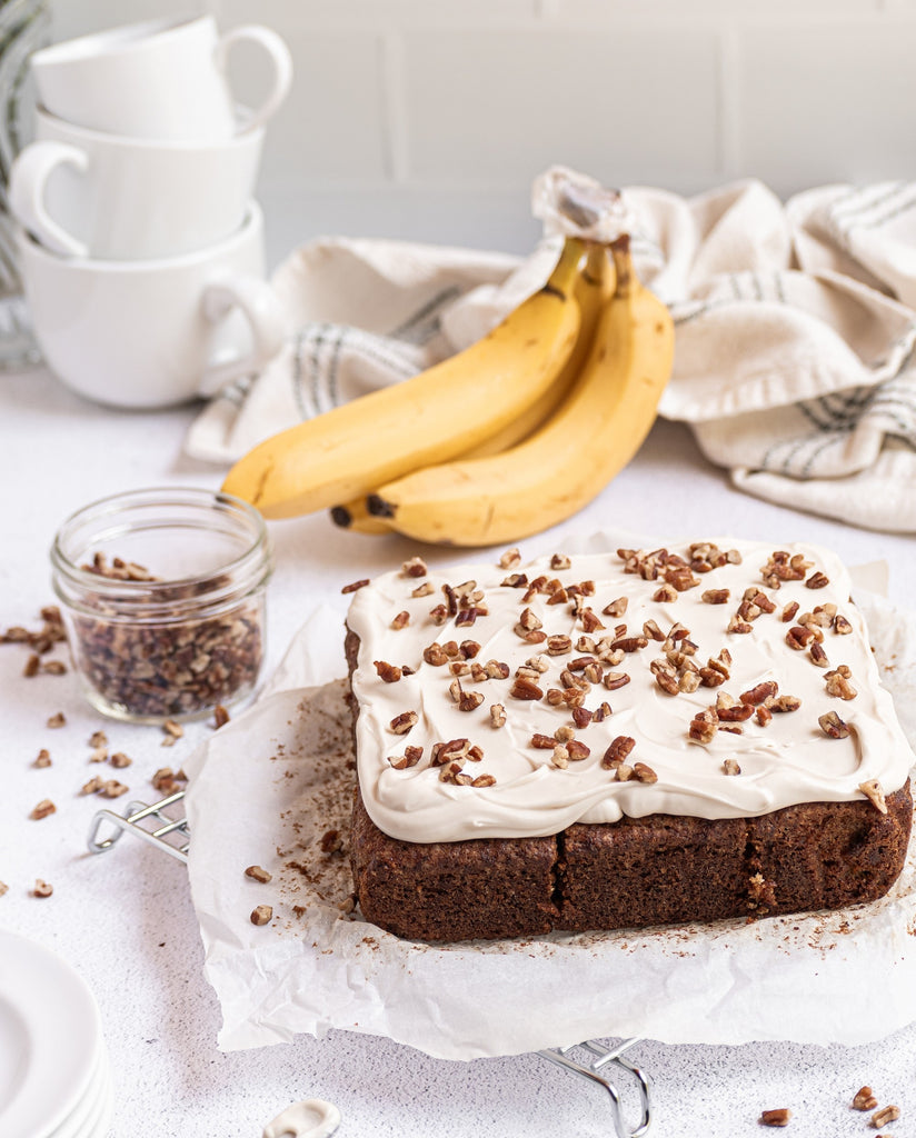 grain-free banana snack cake with frosting and nuts on top. A bunch of bananas, towels, and stacked cups are sitting in background, and a glass of chopped nuts is next to the cake