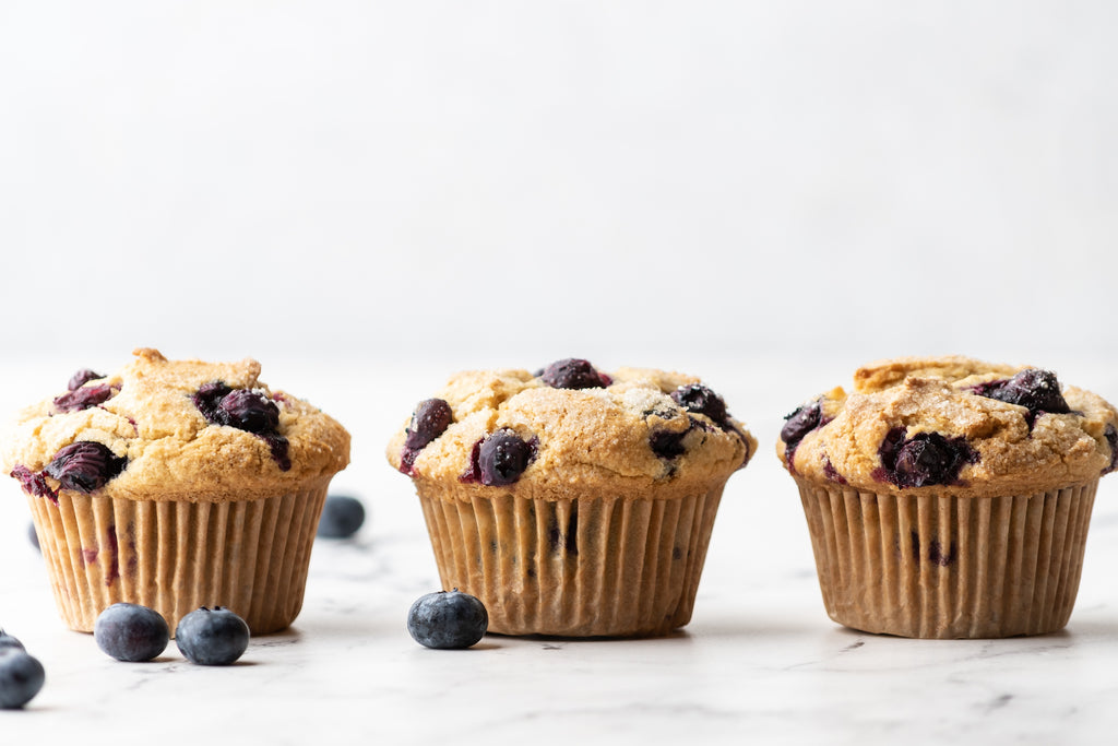 3 blueberry muffins lined up with some blueberries on the counter