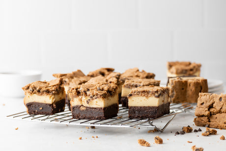 wire rack with squares of grain-free brownie-cheesecake-cookies. Brownie layer is on bottom, cheesecake layer is in middle, and cookie layer is on top.