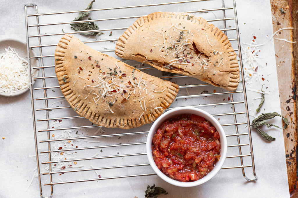 2 grain-free calzones on a wire rack with a bowl of marinara sauce and dried basil