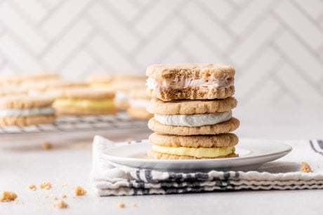 3 sugar cookie sandwiches stacked on a plate. More cookies in the background. Filled with pink, blue, and yellow frosting.