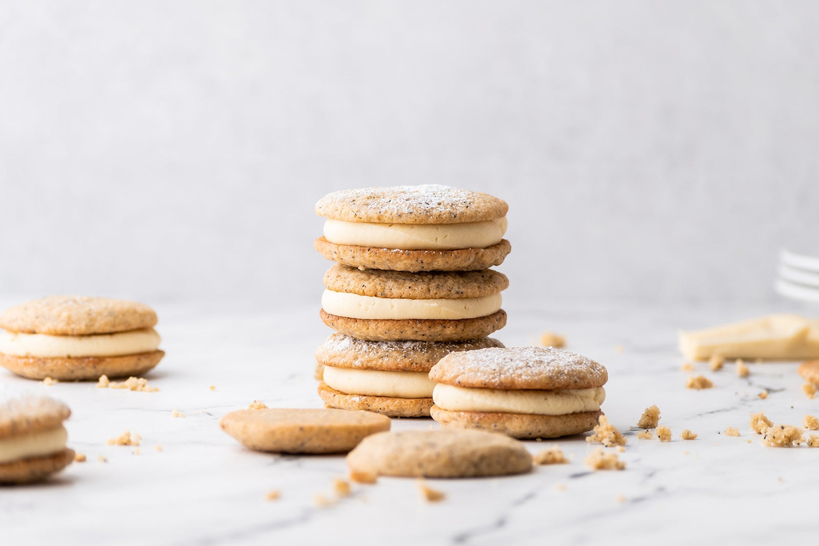 3 lemon poppy seed sandwich cookies stacked, filled with cream cheese icing. More lemon poppy cookies and sandwich cookies sitting nearby on counter