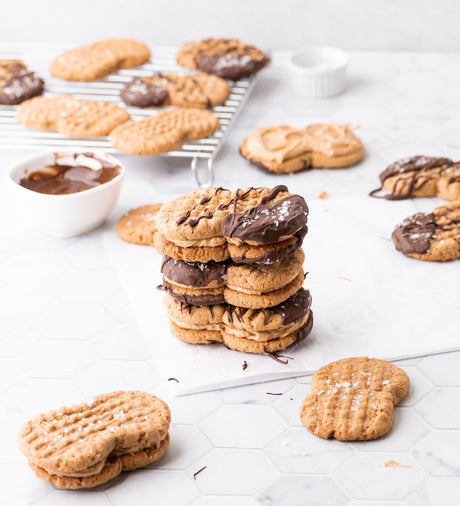 Homemade grain-free peanut-shaped nutter butters. 3 stacked and dipped in chocolate, with more cookies in background on counter.