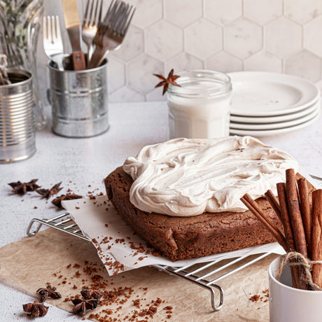 grain-free spice snack cake with frosting sitting on cooling rack. glass of milk, tin of forks, plates, and cinnamon sticks on counter nearby